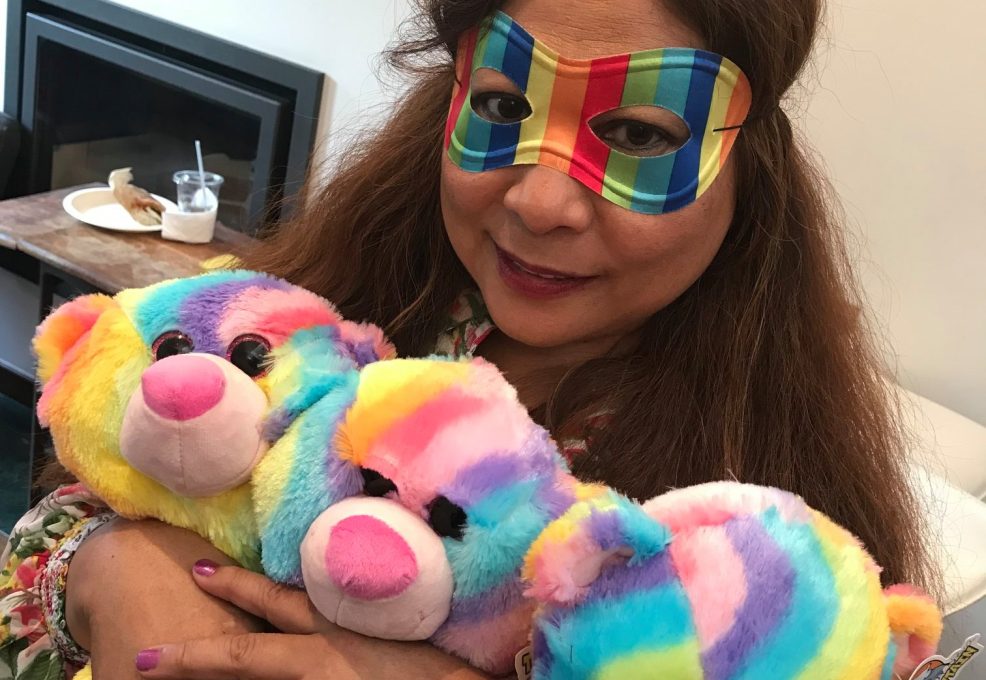A woman dressed festively in a rainbow-colored mask and a mini hat, holding three brightly colored rainbow teddy bears. She has a warm expression as she hugs the bears. The setting appears to be a fun and playful environment, part of a charitable team building event.