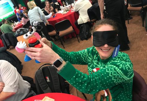 A person wearing a green holiday sweater sits at a table in a large room filled with people. They are holding up a Mr. Potato Head toy while wearing a blindfold, smiling as they participate in an activity. Other attendees in the background are engaged in similar team building holiday-related activities, with festive decorations and red tablecloths creating a cheerful atmosphere.