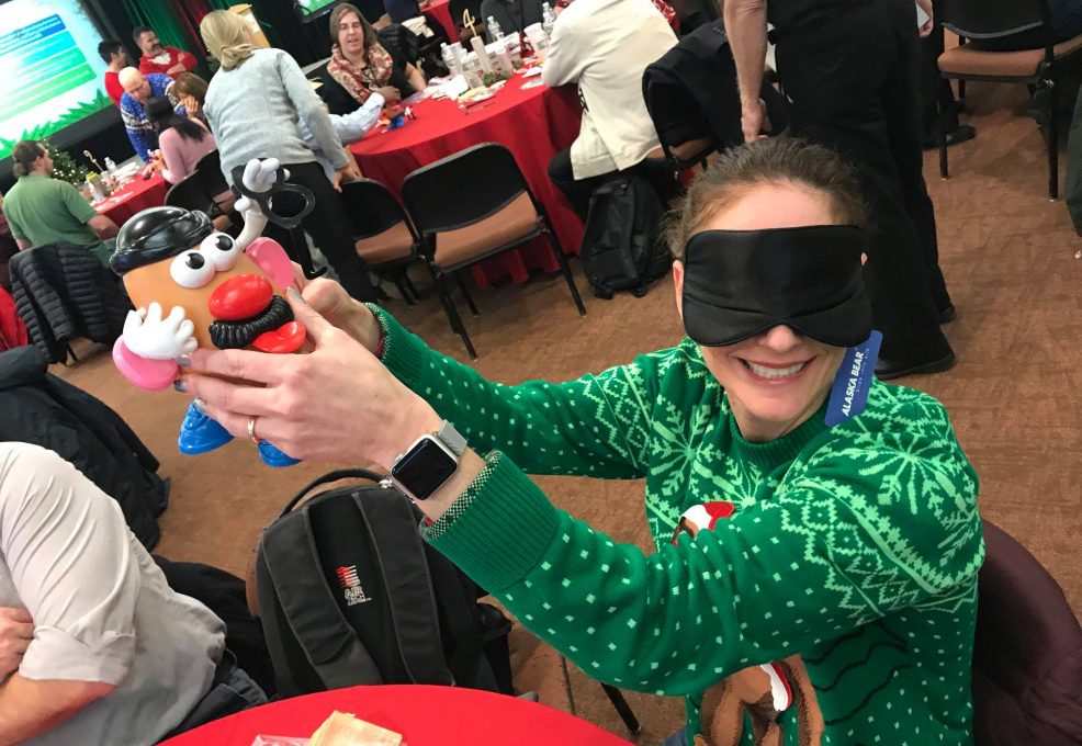 A person wearing a green holiday sweater sits at a table in a large room filled with people. They are holding up a Mr. Potato Head toy while wearing a blindfold, smiling as they participate in an activity. Other attendees in the background are engaged in similar team building holiday-related activities, with festive decorations and red tablecloths creating a cheerful atmosphere.