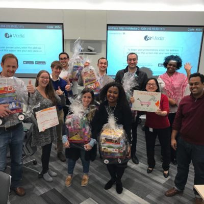 A group of people standing together in a classroom or office setting, smiling and holding gift baskets wrapped in clear plastic. The baskets contain toys, as part of a charitable donation team building event. The group appears to be celebrating their efforts, with some holding signs, and the atmosphere is one of teamwork and achievement. thumbnail