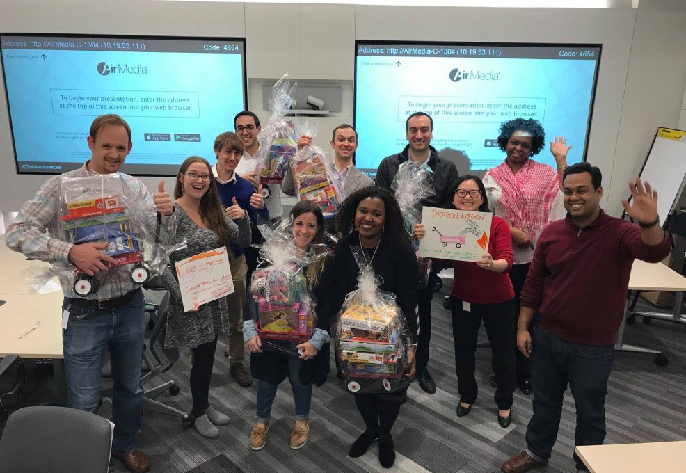 A group of people standing together in a classroom or office setting, smiling and holding gift baskets wrapped in clear plastic. The baskets contain toys, as part of a charitable donation team building event. The group appears to be celebrating their efforts, with some holding signs, and the atmosphere is one of teamwork and achievement.