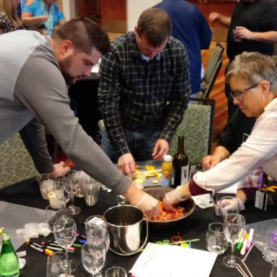 A group of participants actively mixes fruit and ingredients in a large bowl during the 'Winery Team Challenge' event. They are focused on creating their unique sangria blend, using various fruits and drinks on the table. Wine glasses, bottles, and additional supplies are scattered on the table, emphasizing the hands-on nature of the activity. The atmosphere is lively, with other teams visible in the background. thumbnail
