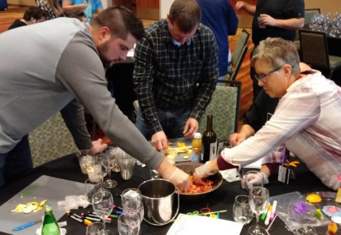 A group of participants actively mixes fruit and ingredients in a large bowl during the 'Winery Team Challenge' event. They are focused on creating their unique sangria blend, using various fruits and drinks on the table. Wine glasses, bottles, and additional supplies are scattered on the table, emphasizing the hands-on nature of the activity. The atmosphere is lively, with other teams visible in the background.