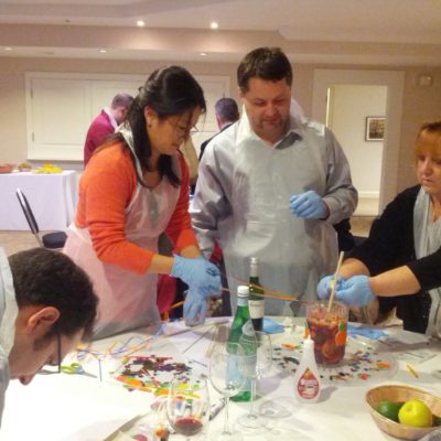 Participants in the 'Winery Team Challenge' work together at a table, wearing aprons and blue gloves as they prepare their signature sangria. The table is covered with colorful decorations, fresh fruit, and bottles, with a pitcher of sangria in the center. The team members appear focused and collaborative, contributing to various aspects of the sangria-making process, from mixing ingredients to adding finishing touches. This image captures the hands-on, creative spirit of the team building event. thumbnail