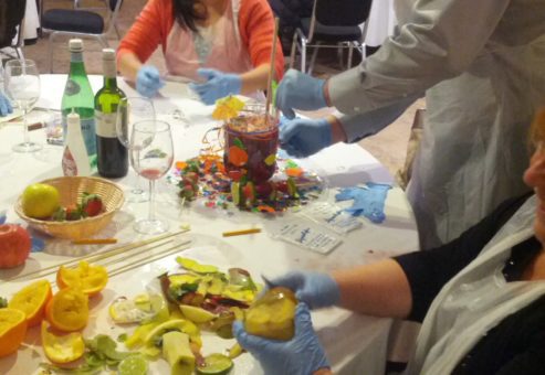 Three participants enthusiastically engage in the 'Winery Team Challenge' team building event. The group is gathered around a table, laughing and preparing their unique sangria blend. One person in the foreground is holding a piece of fruit and wearing blue gloves, while another participant mixes ingredients into a large, decorated sangria pitcher. The table is filled with sliced fruits, wine bottles, and colorful decorations, adding to the fun and creative atmosphere of the event.