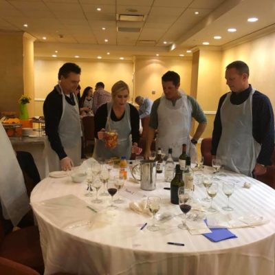 A group of five participants stand around a round table covered with a white tablecloth during the 'Winery Team Challenge.' They are wearing aprons and discussing their next steps as one participant holds a pitcher filled with a colorful sangria blend. The table is scattered with empty wine glasses, bottles of wine, and various ingredients like juices and spices, creating an atmosphere of teamwork and creativity as they finalize their unique sangria creation. thumbnail