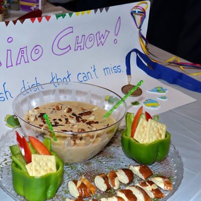 A creatively plated chowder with crackers, fresh vegetables, and a side dish presented at the Wicked Good Chowda Cook-Off team building event, with a sign reading 'Ciao Chow! The fish dish that can't miss.' thumbnail