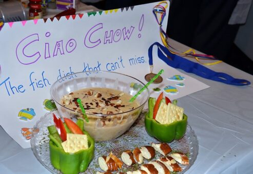 A creatively plated chowder with crackers, fresh vegetables, and a side dish presented at the Wicked Good Chowda Cook-Off team building event, with a sign reading 'Ciao Chow! The fish dish that can't miss.'