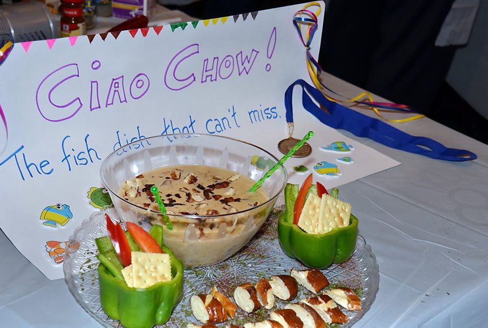 A creatively plated chowder with crackers, fresh vegetables, and a side dish presented at the Wicked Good Chowda Cook-Off team building event, with a sign reading 'Ciao Chow! The fish dish that can't miss.'