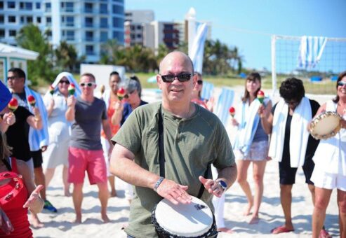Participants engage in a lively Drumming Up A Team event on the beach, using percussion instruments to create a sense of unity and rhythm among the group in a sunny outdoor setting.