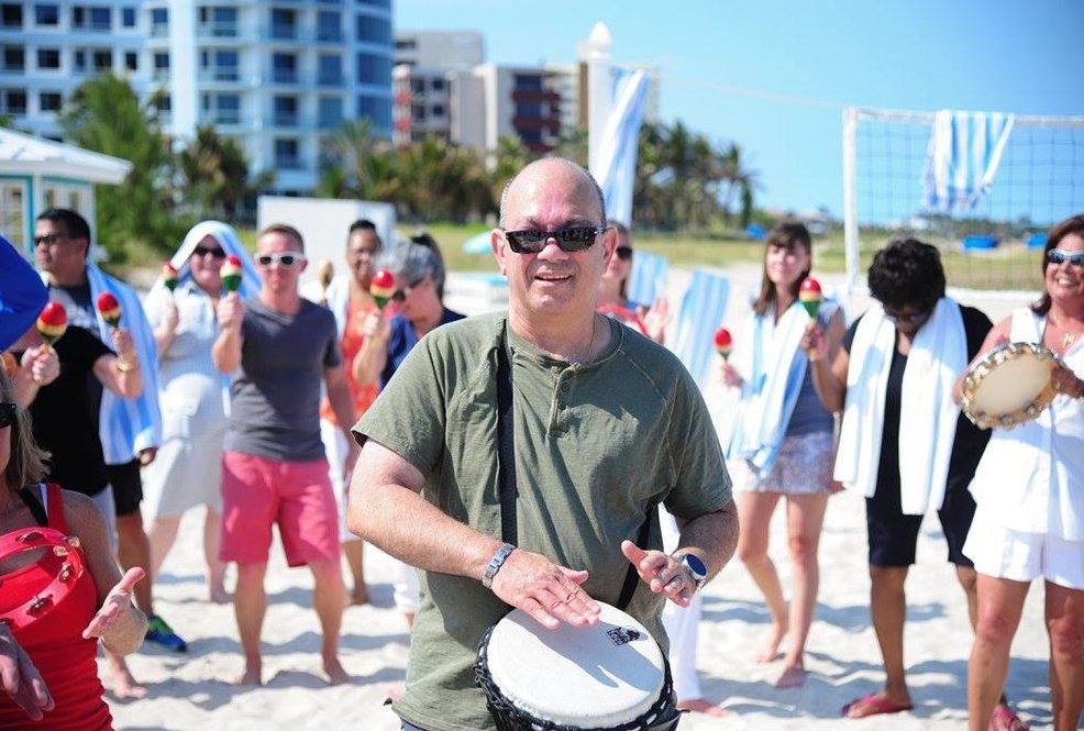 Participants engage in a lively Drumming Up A Team event on the beach, using percussion instruments to create a sense of unity and rhythm among the group in a sunny outdoor setting.