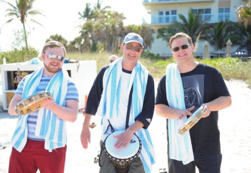 Three participants smiling and drumming together in a Drumming Up A Team event on the beach, wearing striped towels and enjoying the upbeat rhythm in the sunny outdoor setting.