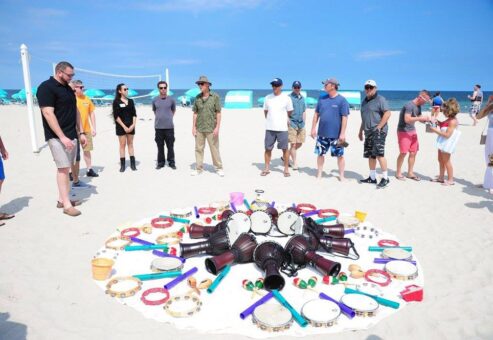 A vibrant array of percussion instruments arranged in a circle on the sand during a Drumming Up A Team event at the beach, with participants standing around preparing to play.