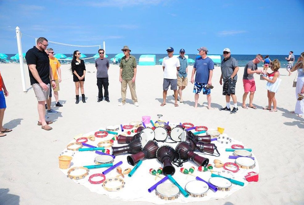 A vibrant array of percussion instruments arranged in a circle on the sand during a Drumming Up A Team event at the beach, with participants standing around preparing to play.