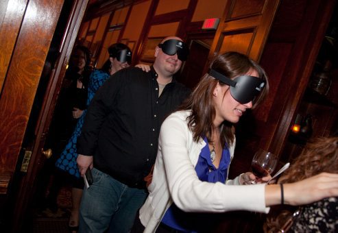 Participants navigating a Dining in the Dark event, wearing blindfolds and engaging with each other while exploring their senses and teamwork during a unique dining experience.