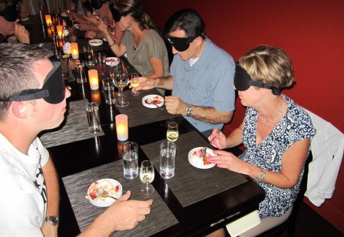 Participants enjoying a meal in a Dining in the Dark experience, with blindfolds on as they engage their senses of taste and smell while sharing the experience.
