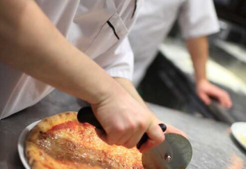A participant cutting a freshly baked pizza with a pizza cutter during The Ultimate Pizza Challenge team building event.