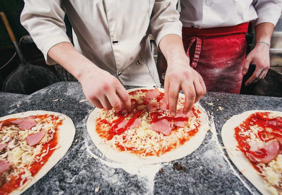 Hands of two participants assembling toppings on pizza dough during The Ultimate Pizza Challenge team building event, focused on culinary teamwork and competition.