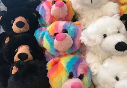 A close-up view of plush teddy bears, showing a variety of colors and designs. Some bears are black with brown accents, while others are brightly colored in a rainbow pattern with pink noses. There are also white polar bear plushies with black noses. The bears are neatly arranged, creating a cheerful, soft display, ready to be customized.