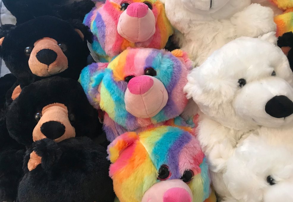 A close-up view of plush teddy bears, showing a variety of colors and designs. Some bears are black with brown accents, while others are brightly colored in a rainbow pattern with pink noses. There are also white polar bear plushies with black noses. The bears are neatly arranged, creating a cheerful, soft display, ready to be customized.