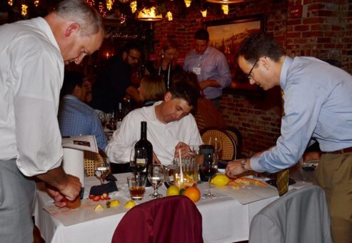 Three participants in the 'Winery Team Challenge' event are focused on preparing ingredients for their sangria creation. Two men are cutting various fruits, such as oranges and lemons, at a table with a bottle of wine, glassware, and other supplies. Another participant seated in the middle is reviewing notes. The event is set in a cozy, warmly lit restaurant with brick walls, as other teams can be seen working in the background, creating a lively and collaborative atmosphere.