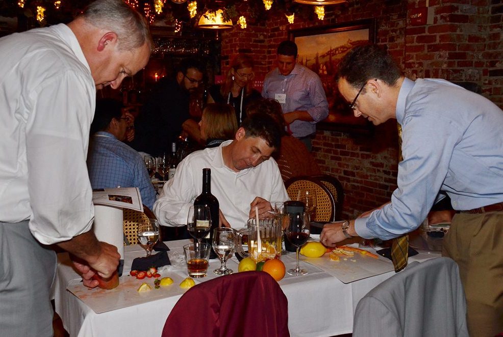 Three participants in the 'Winery Team Challenge' event are focused on preparing ingredients for their sangria creation. Two men are cutting various fruits, such as oranges and lemons, at a table with a bottle of wine, glassware, and other supplies. Another participant seated in the middle is reviewing notes. The event is set in a cozy, warmly lit restaurant with brick walls, as other teams can be seen working in the background, creating a lively and collaborative atmosphere.