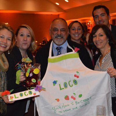 A group of six people smiles and poses for a photo during the 'Winery Team Challenge' event. One person proudly holds a handmade, decorative sign labeled 'LOCO,' while another displays a customized apron with the same name and colorful designs of fruit and flowers. One team member holds a pitcher of sangria garnished with fruit, complete with a fun cocktail umbrella. The atmosphere is warm, with soft lighting in the background, emphasizing the friendly and creative spirit of the team building activity.