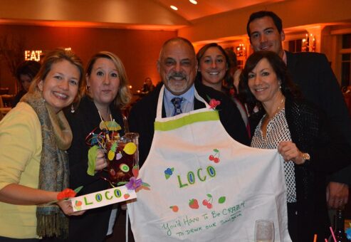 A group of six people smiles and poses for a photo during the 'Winery Team Challenge' event. One person proudly holds a handmade, decorative sign labeled 'LOCO,' while another displays a customized apron with the same name and colorful designs of fruit and flowers. One team member holds a pitcher of sangria garnished with fruit, complete with a fun cocktail umbrella. The atmosphere is warm, with soft lighting in the background, emphasizing the friendly and creative spirit of the team building activity.