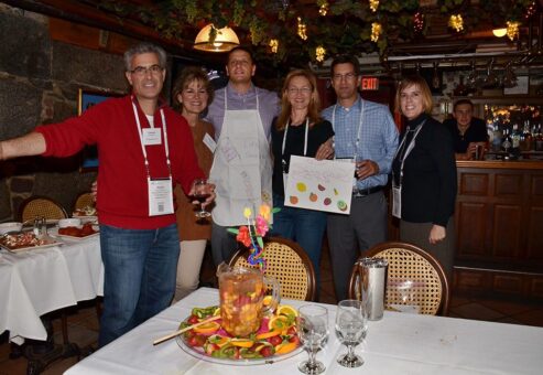 A group of six participants stands together during the 'Winery Team Challenge' event, smiling proudly. They are holding a hand-drawn sign and wearing aprons, standing behind a table with a beautifully decorated pitcher of sangria, surrounded by sliced fruits and garnishes. The cozy restaurant setting features warm lighting and rustic decor. The atmosphere is celebratory, as participants enjoy their creative team building experience.