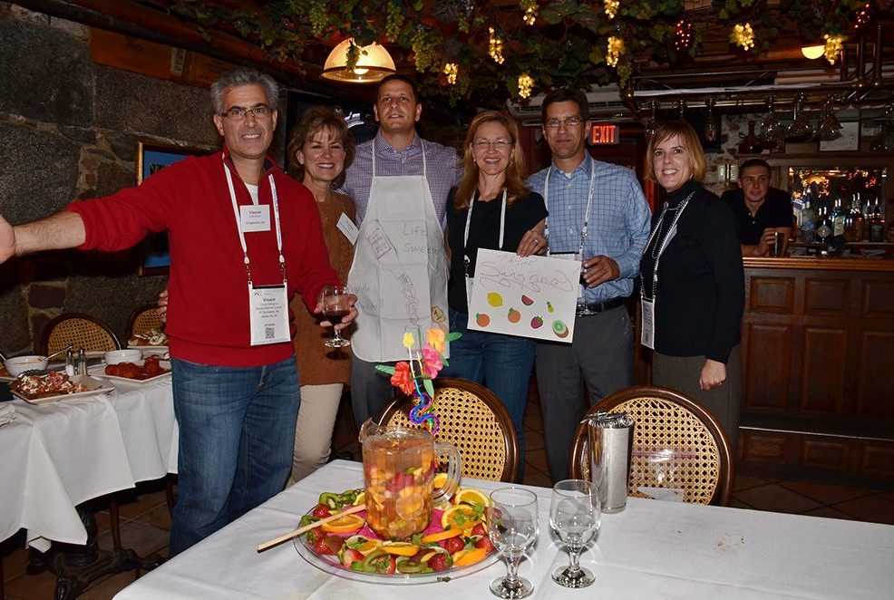A group of six participants stands together during the 'Winery Team Challenge' event, smiling proudly. They are holding a hand-drawn sign and wearing aprons, standing behind a table with a beautifully decorated pitcher of sangria, surrounded by sliced fruits and garnishes. The cozy restaurant setting features warm lighting and rustic decor. The atmosphere is celebratory, as participants enjoy their creative team building experience.
