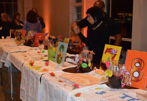 Several colorful sangria presentations are displayed on a long table as part of the 'Winery Team Challenge.' Each sangria pitcher is decorated with vibrant designs, fruit garnishes, and team names or numbers on large cards. The table is covered with personalized aprons, further showcasing each team's creative efforts. In the background, participants are actively engaged in the event, with one person gesturing while others observe. The atmosphere is lively and festive, enhanced by warm lighting.
