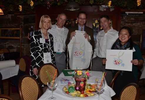 A group of five participants in the 'Winery Team Challenge' event proudly display their team's creations. Two of the members hold up custom-designed aprons and logos for their sangria blend. The group stands around a table adorned with a colorful pitcher of sangria surrounded by decorative fruit and festive drink umbrellas. The setting is a cozy, warmly lit venue with rustic brick walls and grapevine decorations hanging from the ceiling, adding to the wine-themed ambiance.