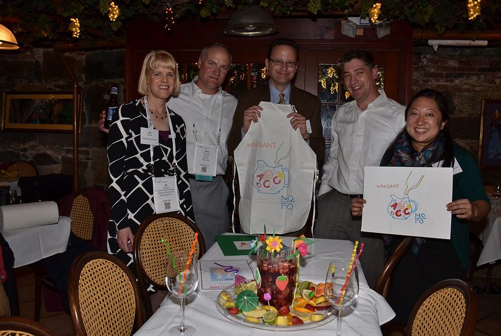 A group of five participants in the 'Winery Team Challenge' event proudly display their team's creations. Two of the members hold up custom-designed aprons and logos for their sangria blend. The group stands around a table adorned with a colorful pitcher of sangria surrounded by decorative fruit and festive drink umbrellas. The setting is a cozy, warmly lit venue with rustic brick walls and grapevine decorations hanging from the ceiling, adding to the wine-themed ambiance.