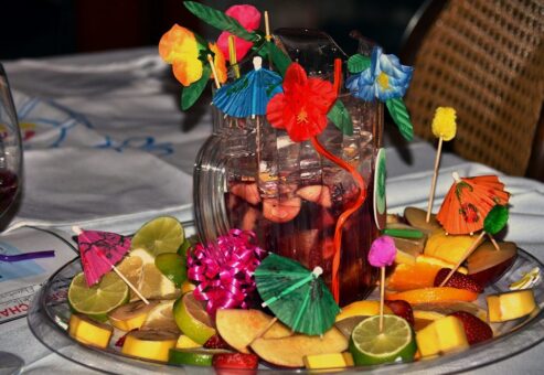 A vibrant pitcher of sangria takes center stage in this presentation from the 'Winery Team Challenge.' The pitcher is decorated with colorful paper umbrellas, flowers, and a festive curly straw. Surrounding the pitcher on a platter are fresh fruit slices, including lime, apple, mango, and strawberries, further adorned with more mini umbrellas and decorative picks. The elaborate arrangement showcases the team's creativity and attention to detail in crafting their signature sangria.