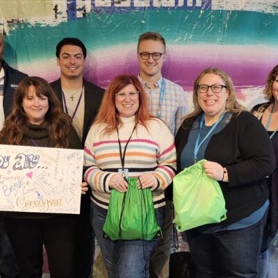 A group of participants smiles while holding colorful drawstring bags filled with care kits during a customized team building event based on the Helping Hands program. One participant holds a sign that reads 'You are amazing!' as they celebrate their contributions. thumbnail