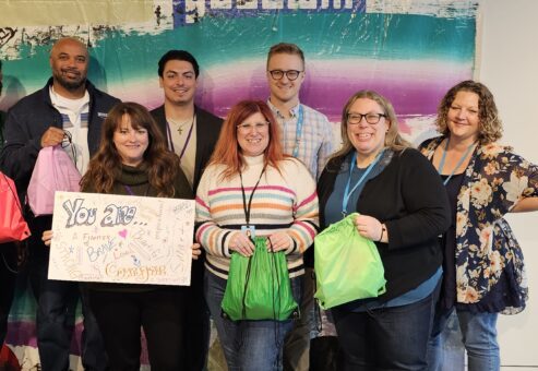 A group of participants smiles while holding colorful drawstring bags filled with care kits during a customized team building event based on the Helping Hands program. One participant holds a sign that reads 'You are amazing!' as they celebrate their contributions.