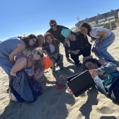 A group of participants poses together on the beach while building a sandcastle during a Custom Team Building event. They are smiling and using a tablet to capture the fun moment in the sun. thumbnail