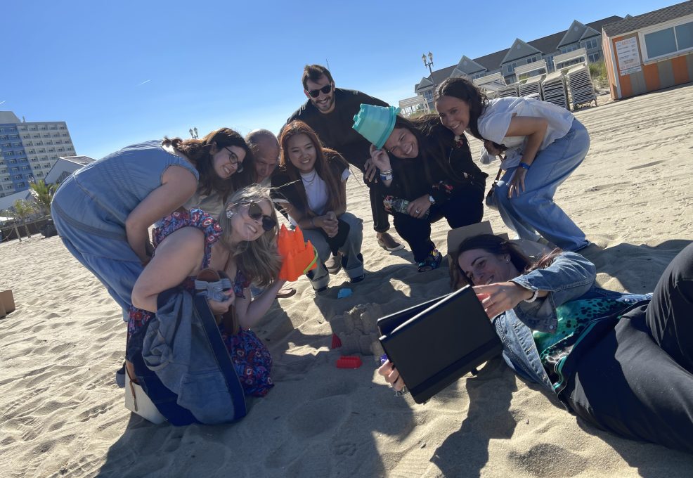 A group of participants poses together on the beach while building a sandcastle during a Custom Team Building event. They are smiling and using a tablet to capture the fun moment in the sun.