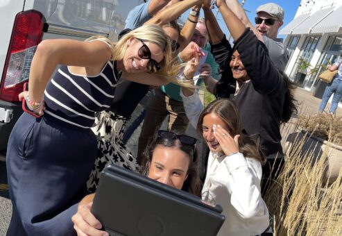 A group of participants huddles together, smiling and giving a high-five while one person takes a selfie with a tablet during a Custom Team Building event. The group is enjoying a fun and energetic moment outdoors.