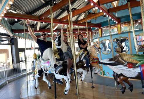 Participants enjoy a ride on a colorful carousel during a Custom Team Building event. They smile and wave as they ride the beautifully crafted horses, adding a playful touch to the experience.
