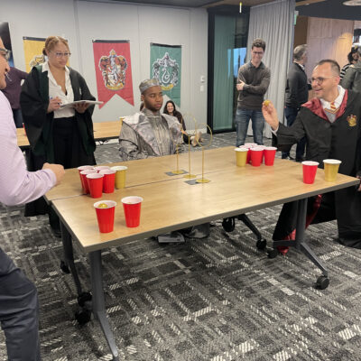 Participants dressed in wizard and knight costumes play a Harry Potter themed game involving cups and a ball during a Custom Team Building event. The room is decorated with banners representing different houses, adding to the magical atmosphere of the activity. thumbnail