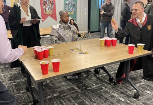 Participants dressed in wizard and knight costumes play a Harry Potter themed game involving cups and a ball during a Custom Team Building event. The room is decorated with banners representing different houses, adding to the magical atmosphere of the activity.