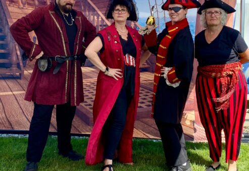 Four participants dressed as pirates pose confidently in front of a pirate ship backdrop during a Custom Team Building event. Their themed attire showcases the creativity and customization of the team building experience.