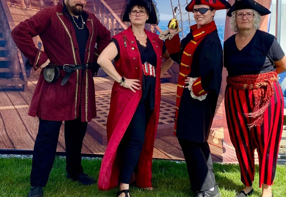 Four participants dressed as pirates pose confidently in front of a pirate ship backdrop during a Custom Team Building event. Their themed attire showcases the creativity and customization of the team building experience.