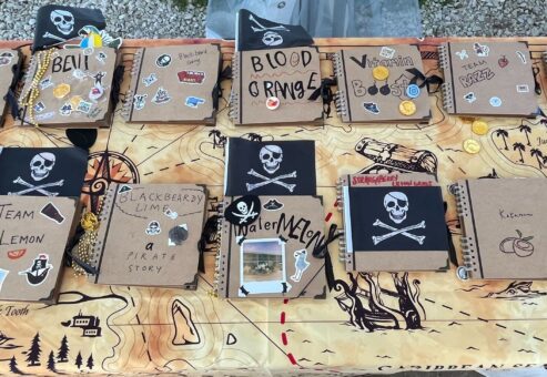 A table filled with pirate-themed notebooks decorated with stickers, skulls, and team names during a Custom Team Building event. The creative decorations reflect the playful pirate theme of the event.
