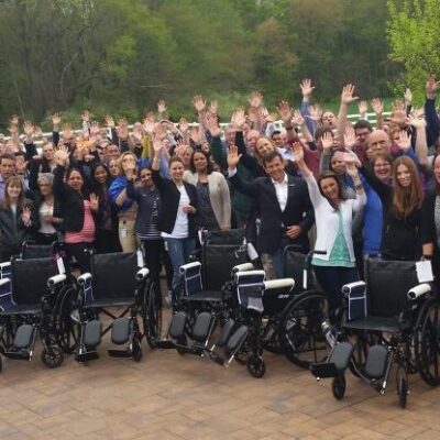A large group of participants gathers outdoors, raising their hands in celebration, standing behind several newly assembled wheelchairs during the Wheelchairs In Motion team building event. The group is smiling and excited, showcasing their completed charitable project. thumbnail