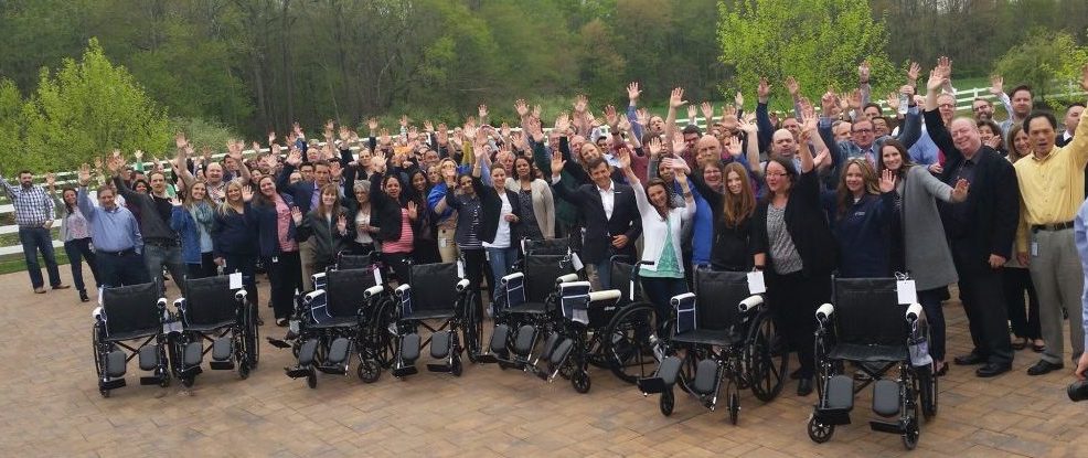 A large group of participants gathers outdoors, raising their hands in celebration, standing behind several newly assembled wheelchairs during the Wheelchairs In Motion team building event. The group is smiling and excited, showcasing their completed charitable project.