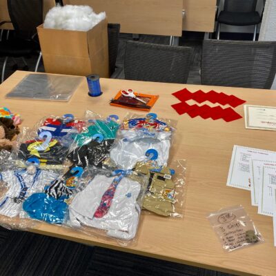 Table setup with teddy bear outfits, materials, and instructions for teams to create stuffed animals as part of The Big Give team building event, where all items will be donated to children in need. thumbnail