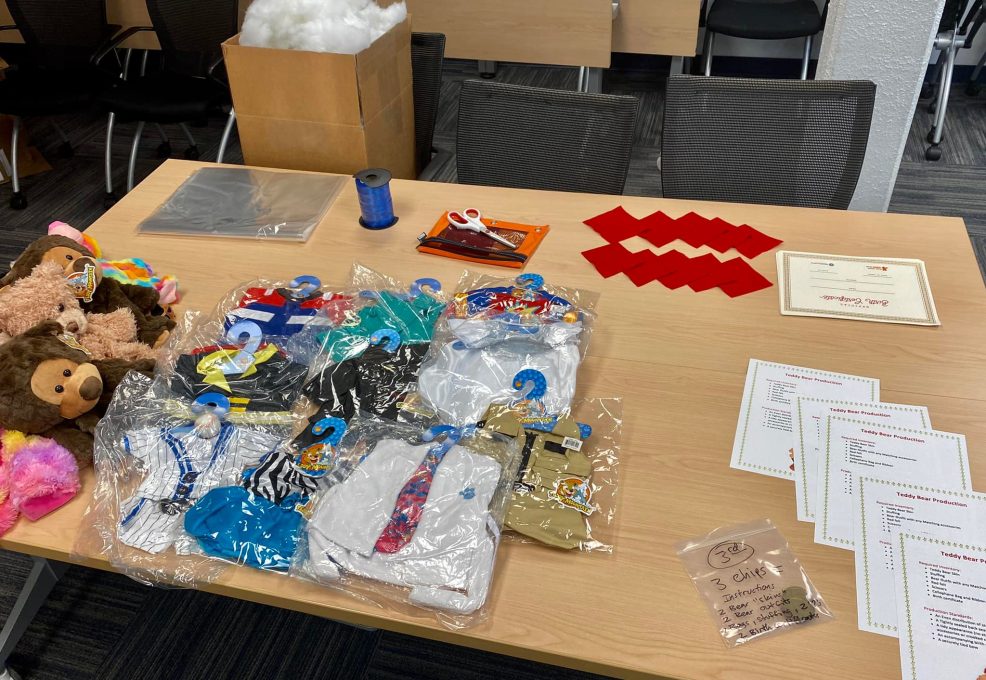 Table setup with teddy bear outfits, materials, and instructions for teams to create stuffed animals as part of The Big Give team building event, where all items will be donated to children in need.