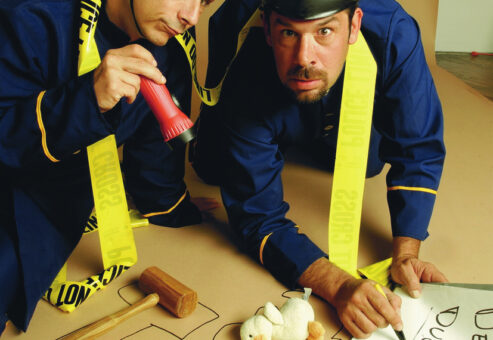 Two actors dressed as police officers humorously investigate a mock crime scene in a playful setup for a Murder Mystery Dinner event. They are kneeling on the ground, surrounded by yellow police line tape, with a chalk outline on the floor resembling the shape of a duck. One officer is holding a red flashlight while the other points at a toy duck with a sign labeled 'Dead Duck.' The scene is lighthearted and comedic, emphasizing the fun, interactive nature of the mystery event.